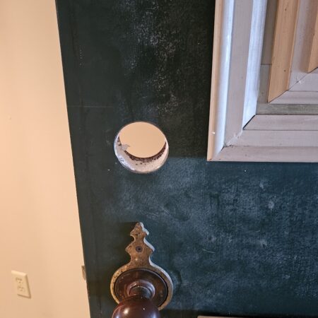 A clean hole drilled into a green wooden door to add a new deadbolt above an existing doorknob.